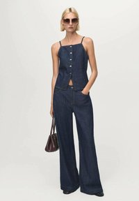 Donkerdenim mouwloze top met knopen en hoge taille brede jeans. Model houdt een bordeauxrode tas vast. Soepele textuur en aansluitend ontwerp.