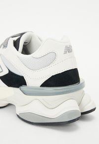 White and black sneaker with mesh and suede materials. Features a chunky sole, layered design, and a translucent accent at the heel.