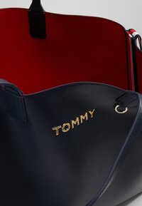 Granatowa skórzana torba na ramię z czerwoną podszewką, z złotym logo "TOMMY" oraz okrąłym detalem w hardware. Gładka tekstura z solidnymi uchwytami.