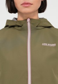 Κοντινό πλάνο ενός ατόμου που φοράει μια πράσινη ζαχαρί ζακέτα με κουκούλα και ροζ φερμουάρ, με το κείμενο «JJXX STUDIO Established in TwentyTwenty» στο στήθος.