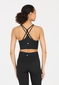 Schwarzes sportliches Croptop mit überkreuzten Trägern auf der Rückseite, gerippter Textur und Logodetail am unteren Rand. Kombiniert mit hochgeschnittenen schwarzen Leggings.