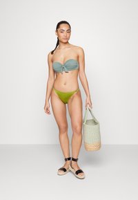 Conjunto de biquíni com um top bandeau verde menta com nó na frente e bottoms verde azeitona, combinado com uma bolsa trançada e sandálias pretas.