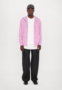 Light pink zip-up hoodie with front pockets, layered over a white t-shirt, paired with black wide-leg cargo pants and black shoes.