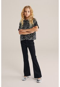 Mustat, säihkemuotoiset lyhythihaiset puserot yhdistettynä mustiin leveälahkeisiin housuihin ja hopeisiin kiiltäviin tasapohjaisiin kenkiin; malli seisoo kädet ristissä.