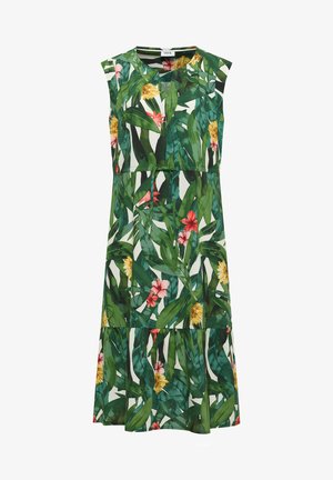Femme portant une robe sans manches, longueur genou, avec un motif de feuilles tropicales vertes et de fleurs rouges, roses et jaunes, debout sur un fond beige.