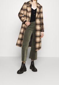 Rutig kappa i brun och beige; svart cardigan under; olivgröna vida byxor; svarta klumpiga ankelstövlar med platå.