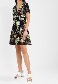 Kvinna iklädd en svart blommig kortärmad kimonoklänning med en utställd kjol och vita öppna klacksandaler med blockklack.