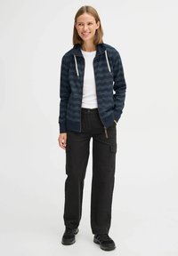 Marinblå zip-up hoodie med vågmönster, dragskohetta och framfickor, i kombination med svarta cargo-byxor och svarta sneakers.