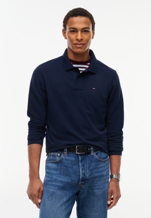 Ung mand iført en marineblå langærmet polo, en stribet undertrøje, blå jeans, sort bælte og et sølvur på håndleddet, stående mod en hvid baggrund.