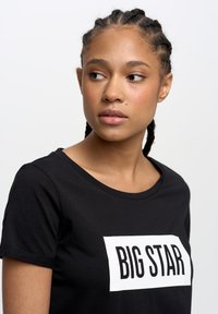 Juodas marškinėliai su apvaliu iškirpimu ir trumpomis rankovėmis, su drąsiu balto stačiakampio spaudiniu, kuris sako "BIG STAR". Lygus audinio paviršius.