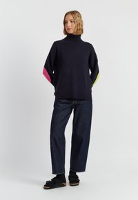 Pull à col roulé oversize de couleur marine avec des accents roses et verts sur les manches, associé à un jean en denim foncé ample et des sandales noires.
