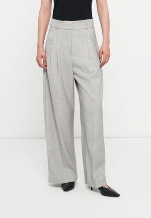 Kobieta nosząca jasnoszare, szerokie spodnie z wysokim stanem, z zakładkami z przodu i czarne czółenka na obcasie z noskiem w szpic.