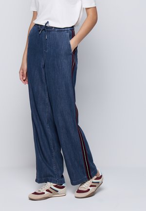 Person, die weit geschnittene Denim-Hosen mit roten und weißen Seitenstreifen, ein weißes T-Shirt und weiße Sneakers mit roten und blauen Akzenten trägt.