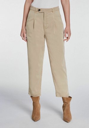 Pantalon taille haute en khaki avec plis sur le devant, jambe droite et ourlets roulés. Fabriqué en tissu doux. Porté avec des bottines en daim marron à mi-mollet.