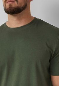 Olijfgroene T-shirt met korte mouwen van katoen en een ronde hals. Bevat een klein, ton-sur-ton logo op de borst. Soepele textuur.