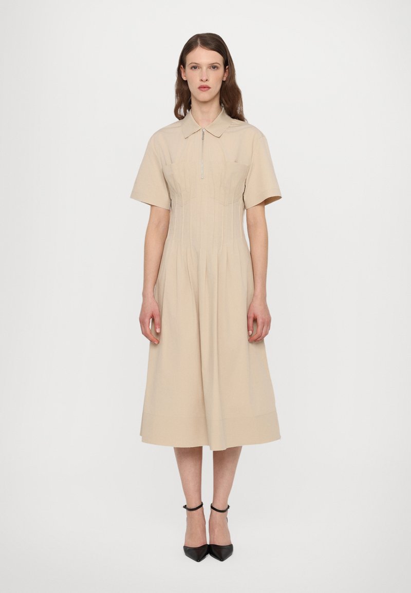 Beige-paittainen mekko, jossa on lyhyet hihat, istuva yläosa, levenevä hame ja edessä vetoketju. Mekko ulottuu juuri polven alapuolelle, ja sen kanssa on mustat korot.