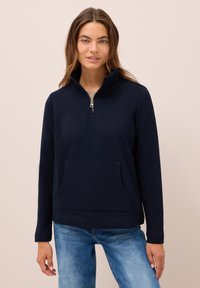 Donna con lunghi capelli castani che indossa una felpa blu navy con zip e jeans blu sbiaditi, su uno sfondo beige semplice.