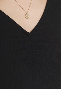 Guldhängehalsband med intrikat mönster som bärs runt halsen över en svart ribbad V-halsplagg.