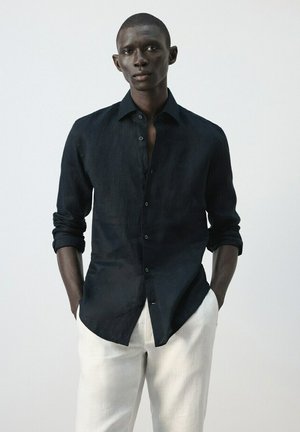 Hombre con camisa oscura de manga larga abotonada y mangas arremangadas, pantalones claros, de pie con las manos en los bolsillos.