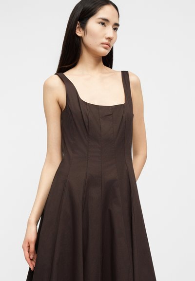 Νεαρή γυναίκα με μακριά σκούρα μαλλιά, που φοράει ένα καφέ αμάνικο φόρεμα με τετράγωνο ντεκολτέ, μπροστά σε φωτεινό απλό φόντο.