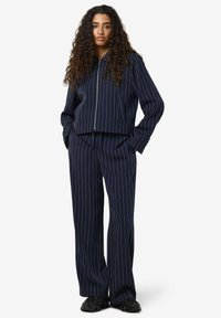 Marineblå dress med pinstripes som inkluderer en kort jakke med glidelås og vide bukser, laget av mykt stoff, kombinert med svarte sko.