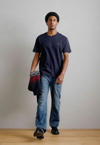 Ναυτικό μπλουζάκι κοντό μανίκι, ανοιχτό μπλε τζιν και μαύρα παπούτσια. Κρατάει μια καρό γκρι και κόκκινη κουβέρτα. Απλή φόντο, ξύλινο πάτωμα.