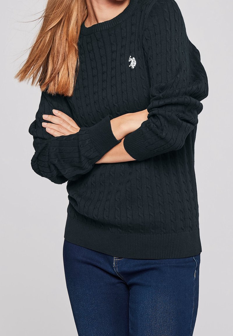Femme aux longs cheveux portant un pull en maille torsadée noir avec un logo blanc brodé et un jean bleu foncé, les bras croisés.
