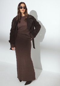 Vestido de punto marrón con un patrón texturizado, combinado con un suéter a juego y un abrigo de trench de ante marrón oscuro. Termina con gafas de sol negras.