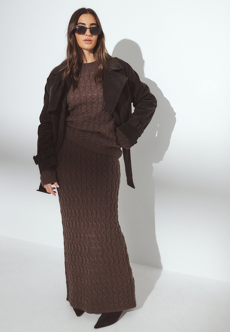 Vestido de punto marrón con un patrón texturizado, combinado con un suéter a juego y un abrigo de trench de ante marrón oscuro. Termina con gafas de sol negras.