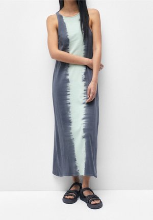 Kobieta w bezrękawnej, sięgającej kostek sukience tie-dye z jasnym paskiem pośrodku i ciemnymi krawędziami, w połączeniu z czarnymi sandałami na sznurki.