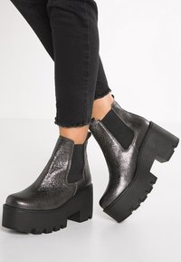 Botas negras hasta el tobillo con un acabado brillante y texturizado, que cuentan con paneles laterales elásticos y una suela de plataforma gruesa con un diseño con relieve.