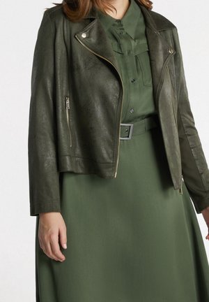 Femme portant une robe boutonnée vert olive foncé avec une large ceinture et une veste en cuir vert foncé courte avec des fermetures éclair argentées.
