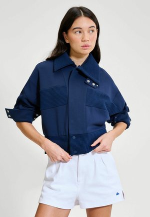 Kobieta w granatowej, teksturowanej kurtce z podwiniętymi rękawami i białych krótkich spodenkach, stojąca na jasnym, jednolitym tle.