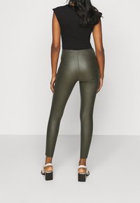 Leggings de cuero sintético verde oliva con cintura alta, diseño ajustado y textura suave. Combinados con una blusa negra de mangas cortas y sandalias blancas de tacón.