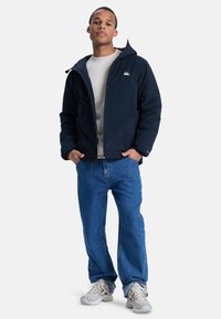 Marine vandtæt jakke med hætte, frontlynlås og logo på brystet, båret over en let skjorte og parret med blå jeans og grå sneakers.