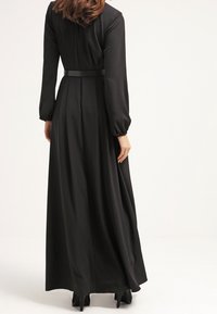 Robe noire longue à manches longues, corsage ajusté et ceinture cintrée à la taille. Le tissu lisse tombe jusqu'au sol.