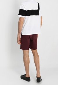 Bílý tričko s černým horizontálním pruhem na zádech, spojené s kaštanovými šortkami a černými teniskami. Ležérní střih, bavlněný materiál.