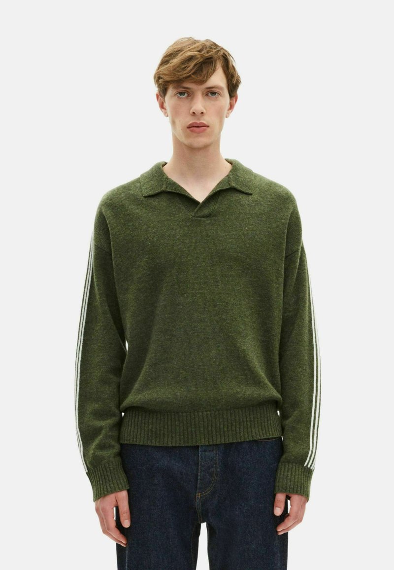 Jeune homme portant un pull en tricot vert avec des rayures blanches sur les manches et un jean bleu foncé, se tenant contre un fond uni.