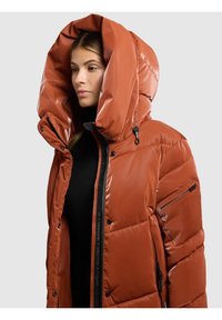 Chaqueta acolchada naranja con una gran capucha, textura brillante, cierre frontal con cremallera y bolsillos laterales con cremallera. Cuenta con botones de presión en el cuello.