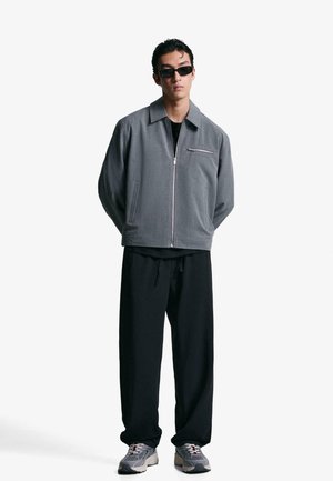 Sivi zip-up jakna s ravnim ovratnikom, nosi se preko crne majice. Labave crne hlače i sivi tenisice. Minimalistički dizajn s čistim linijama.
