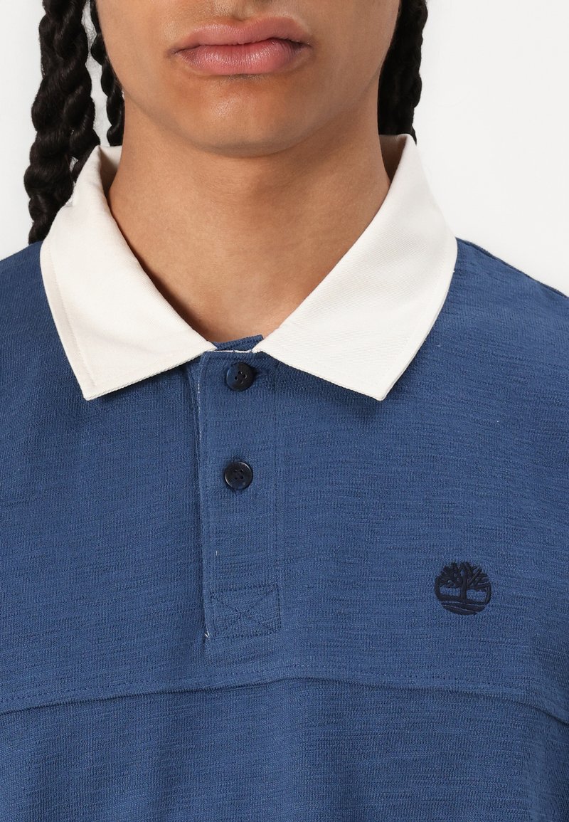 Polo shirt bleu avec une finition texturée, col blanc et trois boutons. Présente un logo noir brodé sur la poitrine.