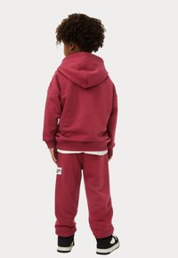 Bordeaux hoodie met bijpassende joggers, met een voorzak en een wit logo-embleem, gecombineerd met zwarte sneakers.