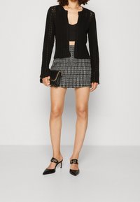 Czarny dziergany kardigan z otwartym przodem i prążkowanymi detalami, zestawiony z dopasowaną spódnicą w monochromatyczną kratę. Czarne szpilki z szpiczastym noskiem i złotymi akcentami.