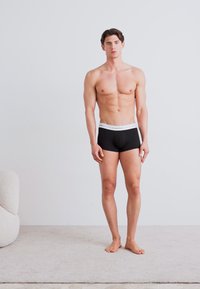 Schwarze Boxerbriefe mit einem kontrastierenden weißen Logo-Bund, der Stoff wirkt glatt und dehnbar, getragen von einem stehenden männlichen Modell in einer neutralen Umgebung.