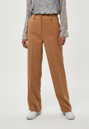 Femme portant un pantalon camel taille haute à jambes larges, associé à des baskets blanches et un haut bleu à fleurs à manches longues.