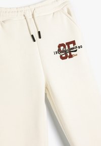 Cremefarbene Sweatpants mit verstellbarem Kordelzug, die einen grafischen Print in Burgunderrot und Grün zeigen, der "California 1980" und "SF State" beinhaltet.