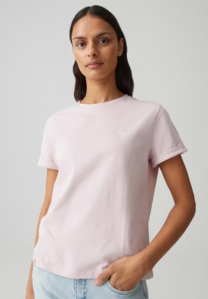 Vrouw met lang donker haar draagt een lichtroze T-shirt met korte mouwen en een klein hartembleem, lichtblauwe jeans, staand tegen een effen achtergrond.