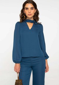 Chemisier bleu sarcelle avec un col en V et des épaules froncées, doté de manches longues et bouffantes. Tissu lisse et léger avec peu de détails de conception.