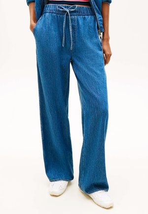 Persoon draagt losse blauwe spijkerbroek met elastische tailleband en trekkoord, gecombineerd met witte sneakers en handen in de zakken.