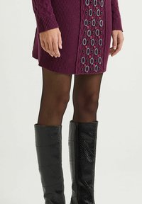 Robe tricotée magenta avec des détails à motifs en bleu et bordeaux. Associée à des bottes noires texturées montant jusqu'aux genoux.